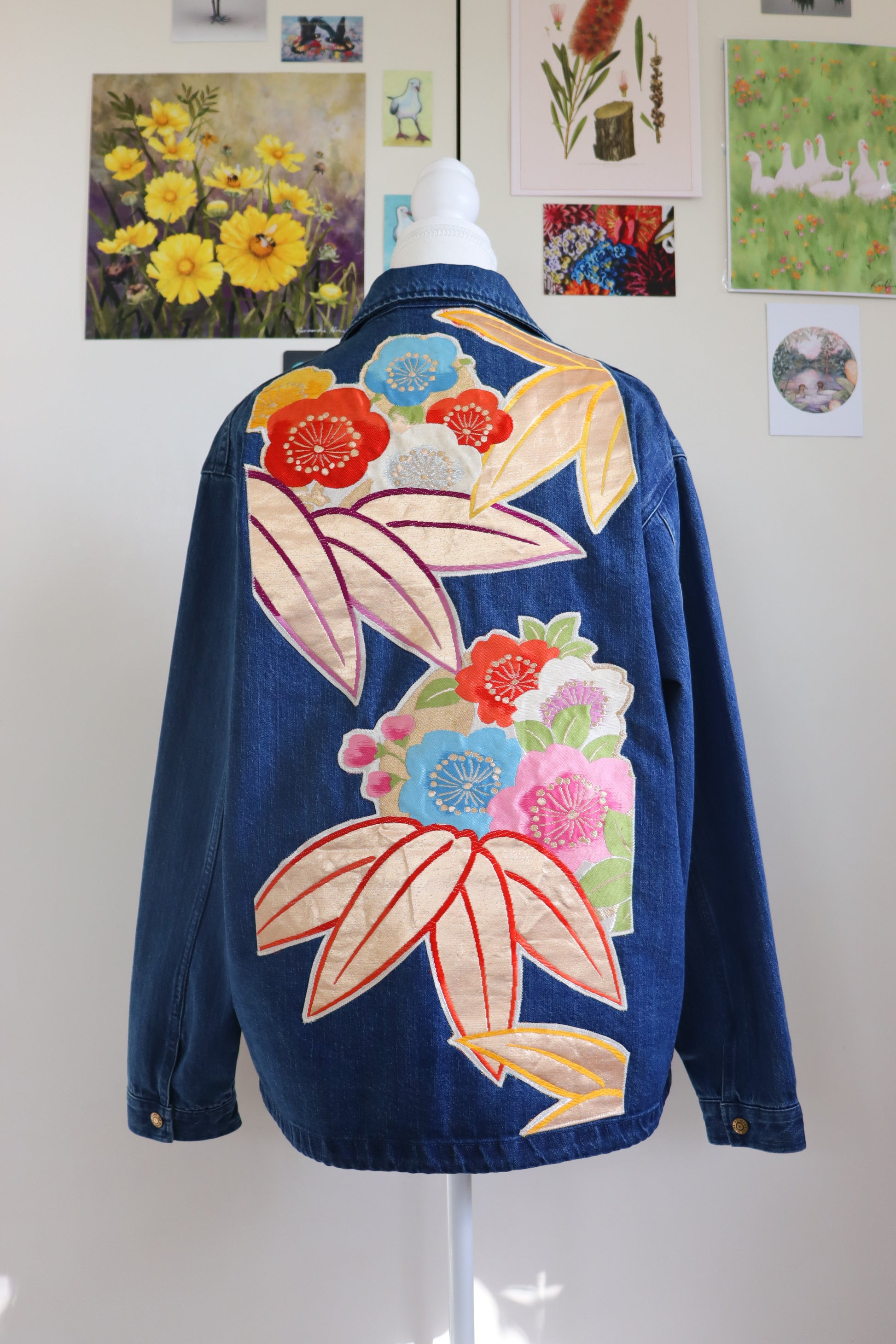 Blue denim jacket with colorful floral embroidery on a mannequin against a white wall with floral prints.