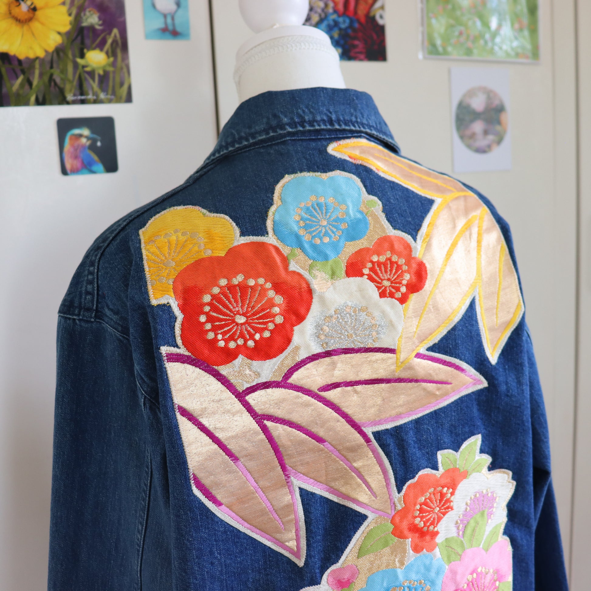 Denim jacket with colorful floral embroidery on a mannequin against a wall with framed pictures.