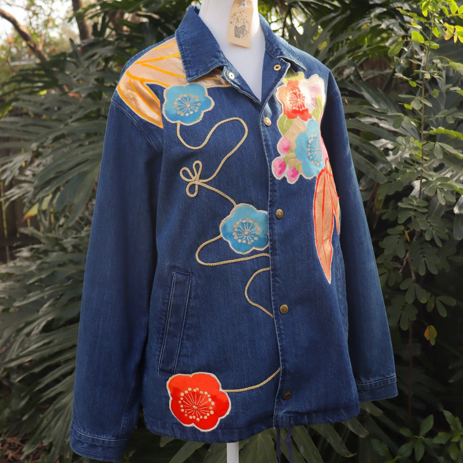 Blue denim jacket with colorful floral embroidery on a mannequin against a natural background.