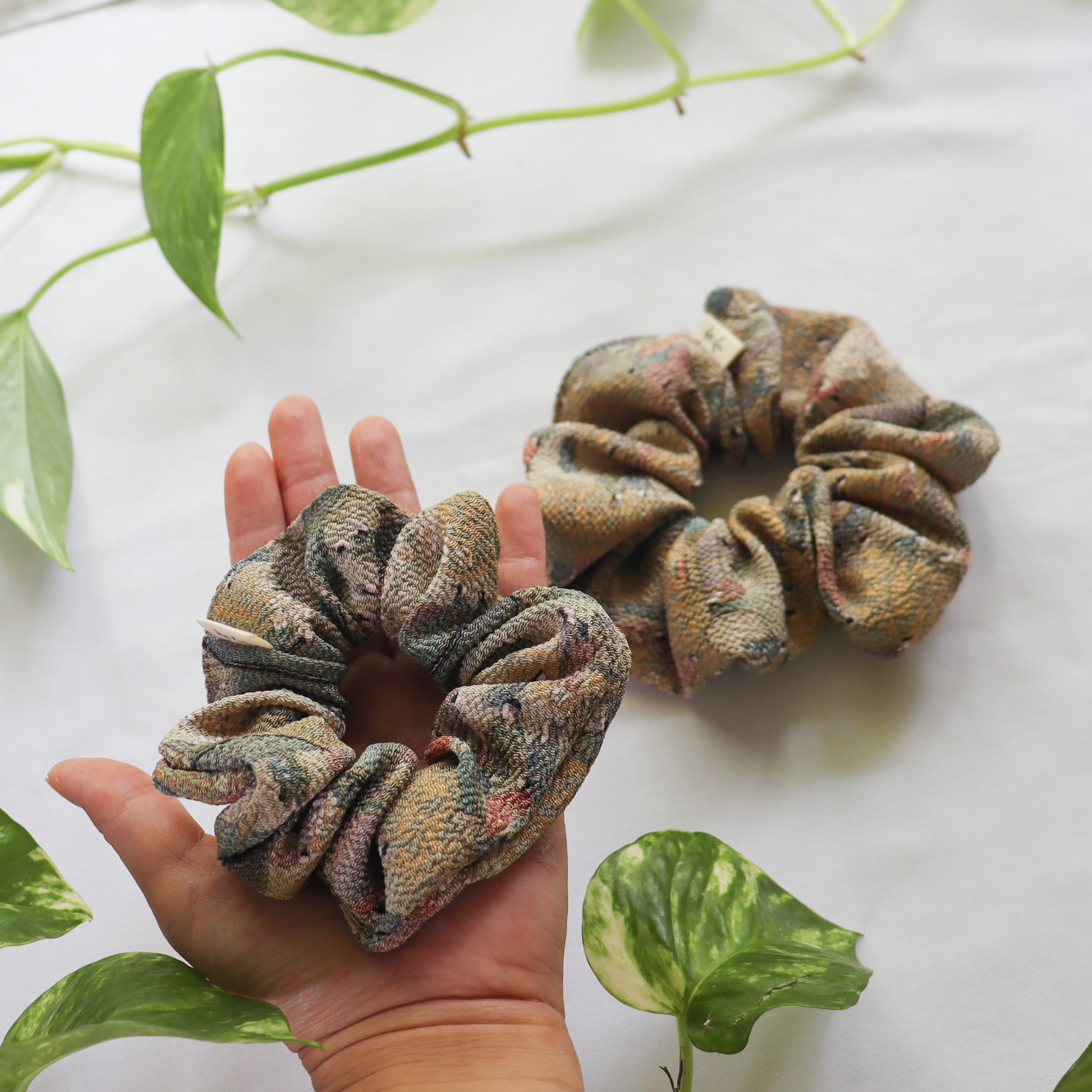 Two patterned scrunchies on a hand with green leaves in the background