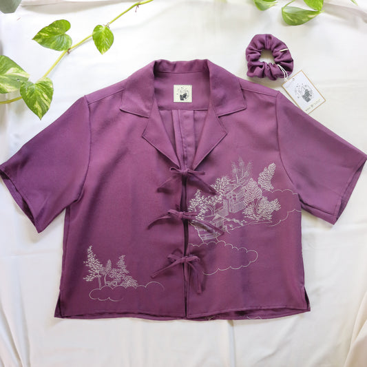 Purple blouse with floral pattern on a white surface with green leaves.